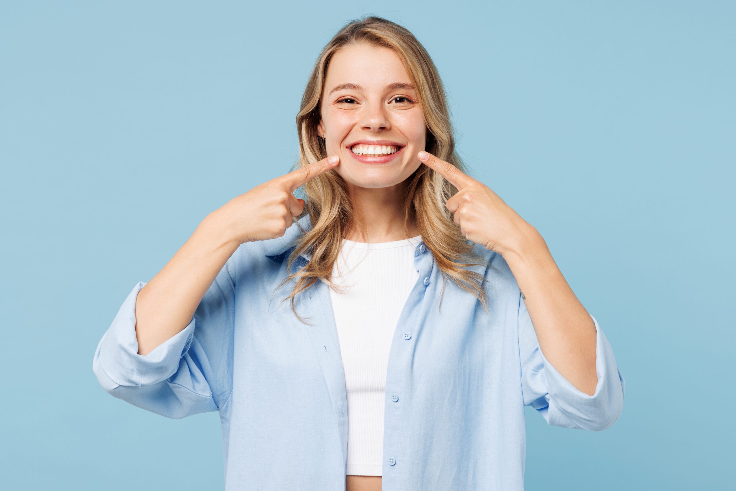 Young smiling woman pointing index fingers to opened mouth, showing professionally whitening teeth
